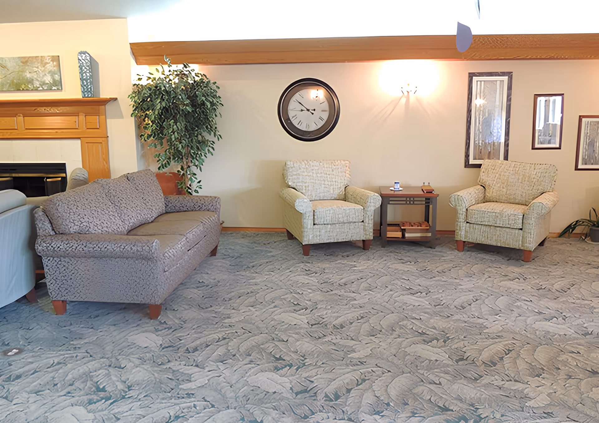 A cozy living room area with a patterned carpet, a gray sofa, two beige armchairs, a wooden side table with books and small items, a large wall clock, framed artwork, a potted plant, and a wooden fireplace mantel.
