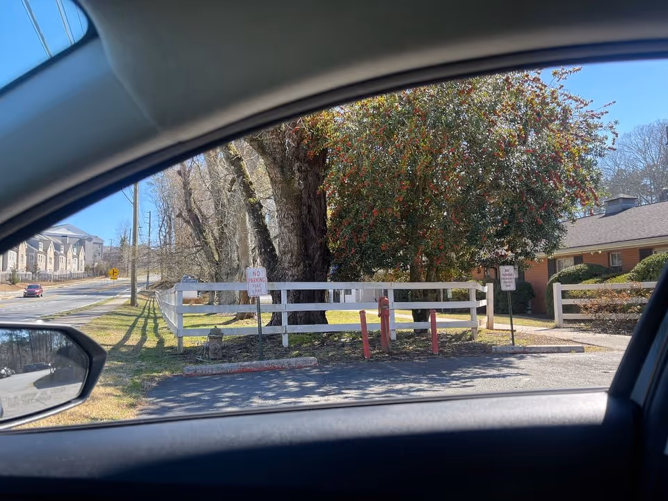 View from inside a car through the side window showing a white fence, a large tree with berries, and the front of a single-story brick building set behind a lawn.