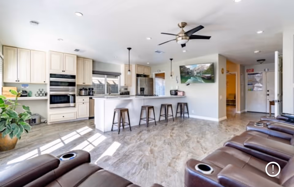 Bright and spacious open-concept room featuring a modern kitchen with cream-colored cabinets, stainless steel appliances, and a large island with four bar stools. The room also includes a row of brown leather recliners with cup holders, a ceiling fan, a large plant near the window, and a wall-mounted TV displaying a nature scene. The floor is covered with light-colored tiles, and there is a hallway leading to other areas of the facility.