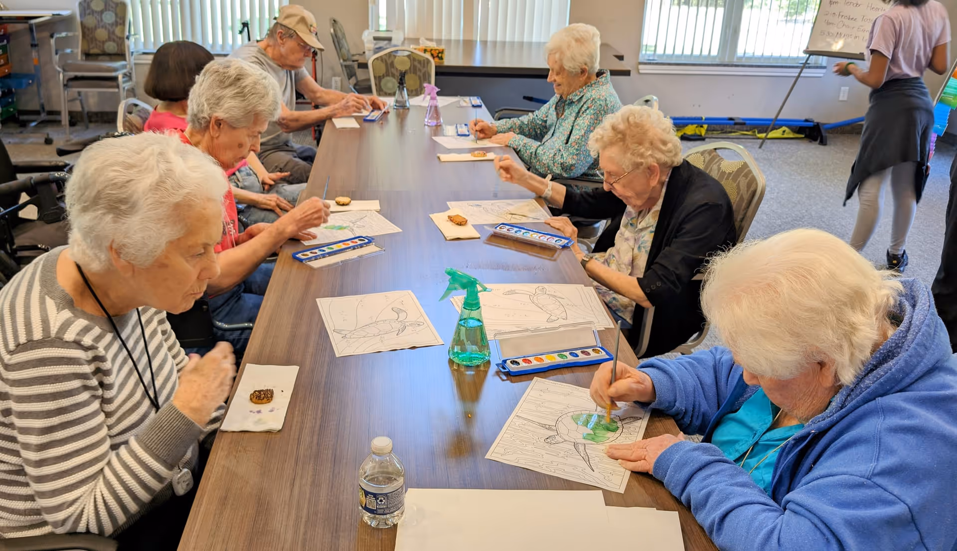 A group of elderly individuals seated around a long table engaged in a watercolor painting activity. Each person has a paint set and a coloring sheet with a bird design. There are snacks and a water bottle on the table. The room has large windows with vertical blinds and a whiteboard in the background.