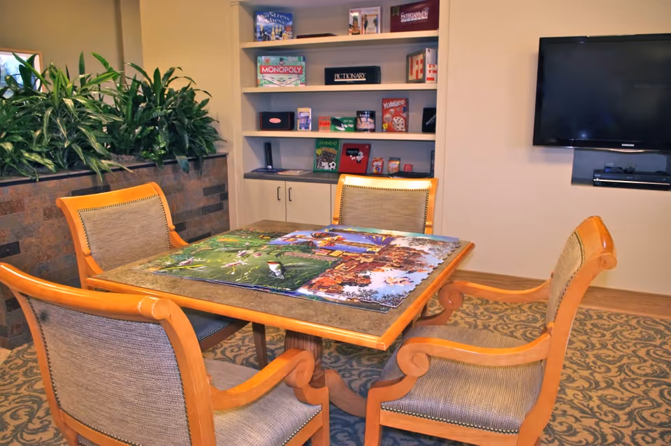 A cozy game room with a square wooden table and four cushioned chairs arranged around it. A partially completed jigsaw puzzle is on the table. Behind the table is a built-in shelf filled with board games like Monopoly, Pictionary, and Yahtzee. To the right, a flat-screen TV is mounted on the wall. There are green plants on a ledge to the left and a patterned carpet on the floor.