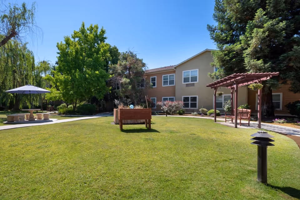 Sunny landscaped courtyard with a green lawn, benches, pergola, umbrella-covered table, and a two-story residential building in the background.
