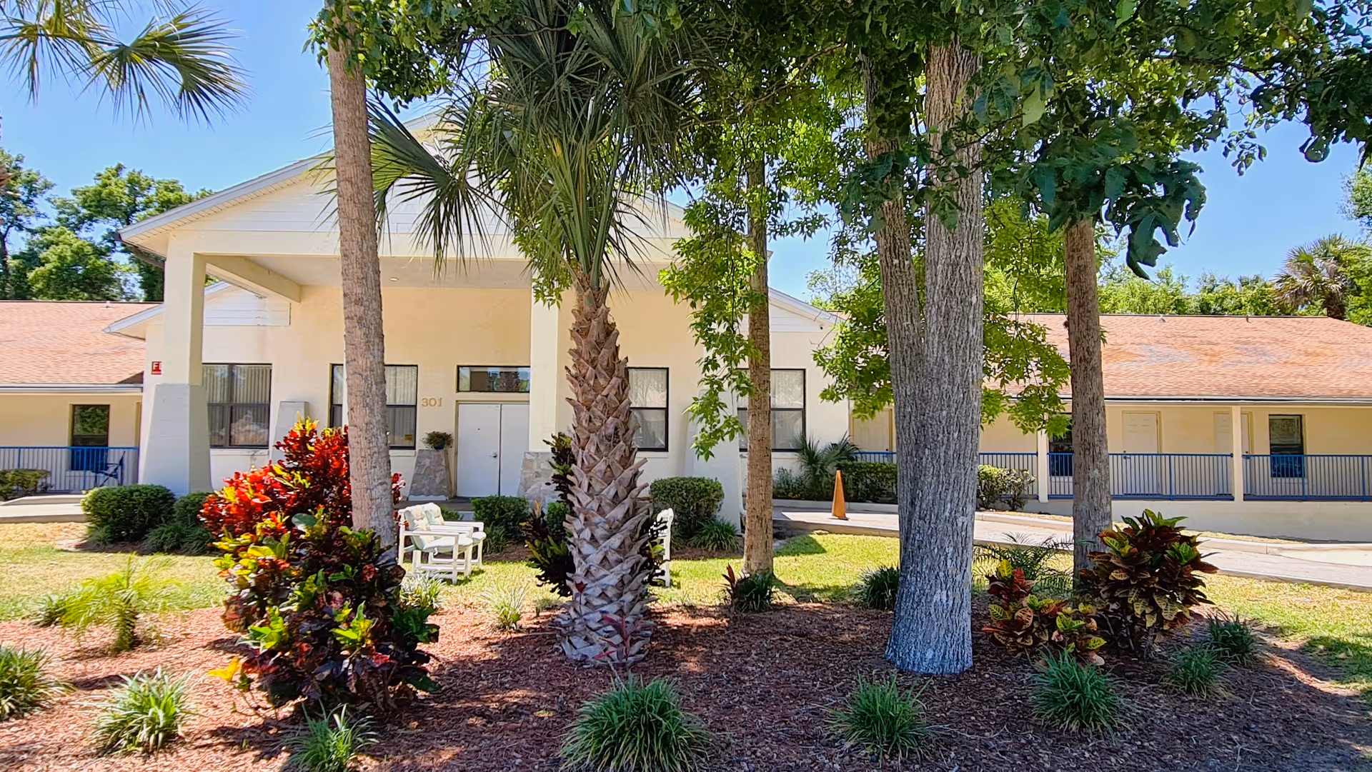 Exterior view of Crown Assisted Living facility showing a single-story building with a covered entrance, surrounded by palm trees, other trees, and landscaped bushes under a clear blue sky.