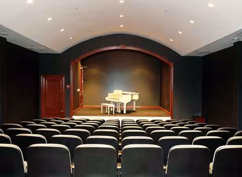 An empty auditorium with rows of seats facing a small stage and piano.