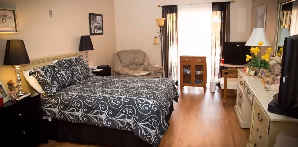 A cozy bedroom with a bed covered in a black and white patterned bedspread, two bedside tables with lamps, a beige armchair, a wooden cabinet near a window with sheer curtains, and a dresser with a TV, lamp, flowers, and framed photos.