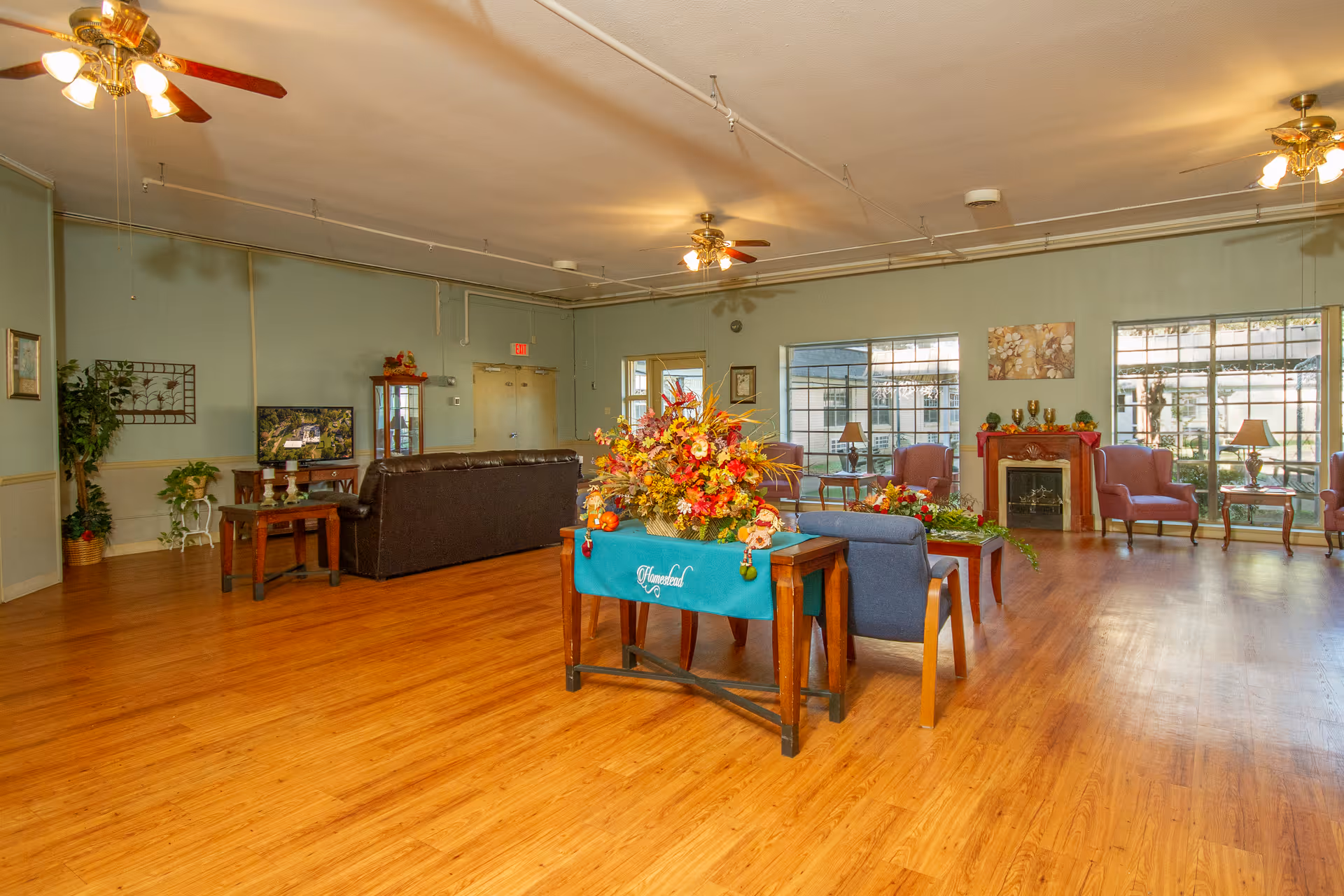 Spacious senior living common room with seating areas, hardwood floors, ceiling fans, large windows and a central table topped with a floral arrangement.