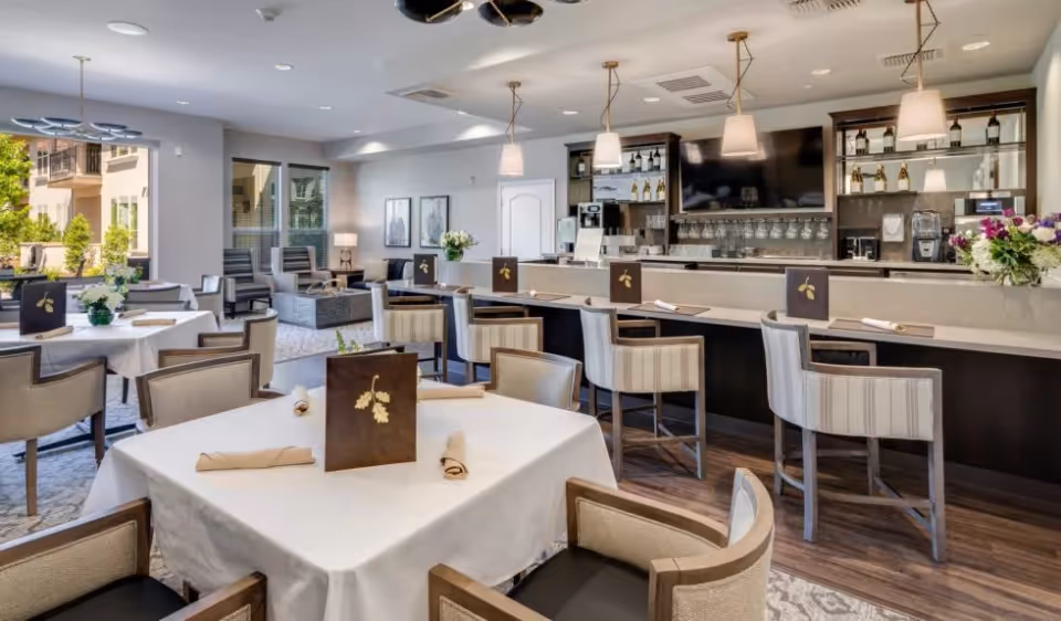 Bright dining room and bar area with set tables, upholstered chairs, and high stools in a communal living space.