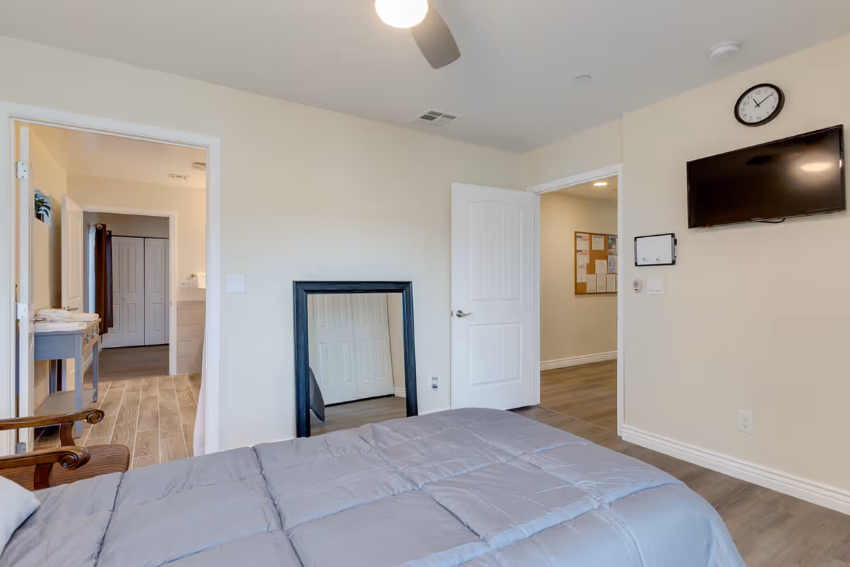 A bedroom with a bed covered in a gray comforter, a wooden chair, a large mirror leaning against the wall, an open door leading to a hallway with a bulletin board, and another open door showing a bathroom area with a sink and closet doors. A wall-mounted TV and a clock are visible above the bed.