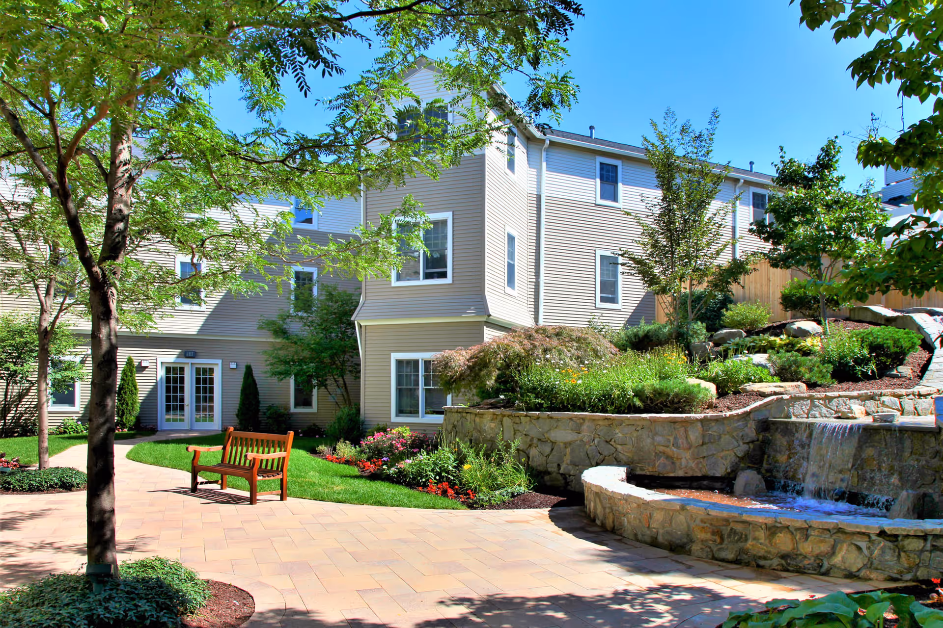 Outdoor courtyard area of a senior living facility with a stone water fountain, landscaped garden beds, trees, a wooden bench, and a multi-story beige building in the background under a clear blue sky.