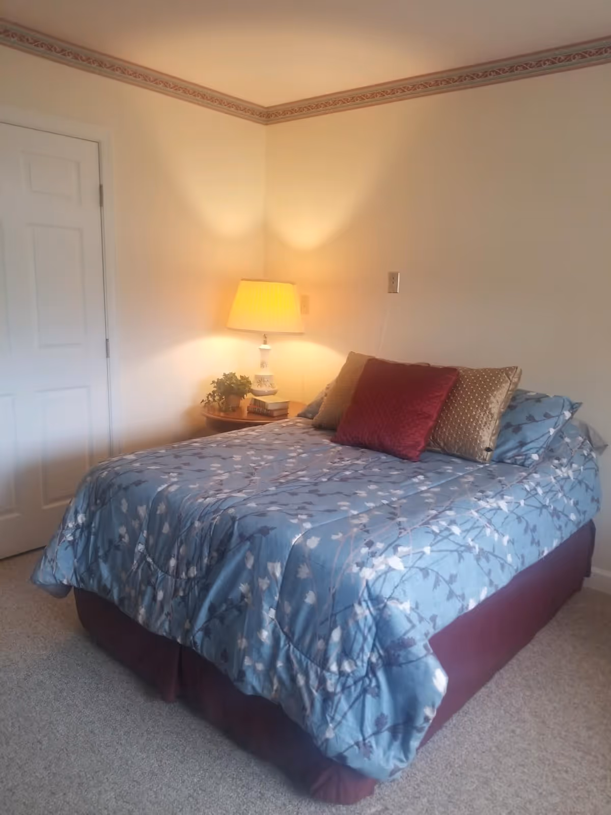 A bedroom with a bed covered in a blue floral comforter and three decorative pillows in red and gold. There is a wooden nightstand next to the bed with a white lamp, a small plant, and some books. The walls are light-colored with a decorative border near the ceiling, and a closed white door is visible on the left side.