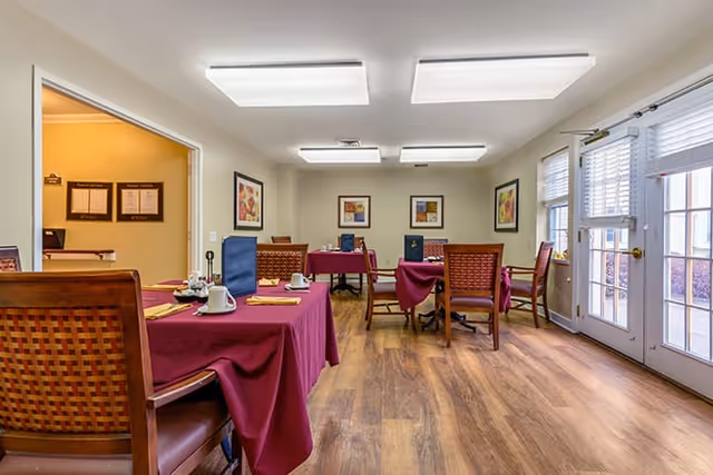 A dining room with wooden floors and tables covered with burgundy tablecloths. Each table has place settings with cups, napkins, and menus. The room is well-lit with ceiling lights and has framed artwork on the walls. There are large windows and a glass door letting in natural light.