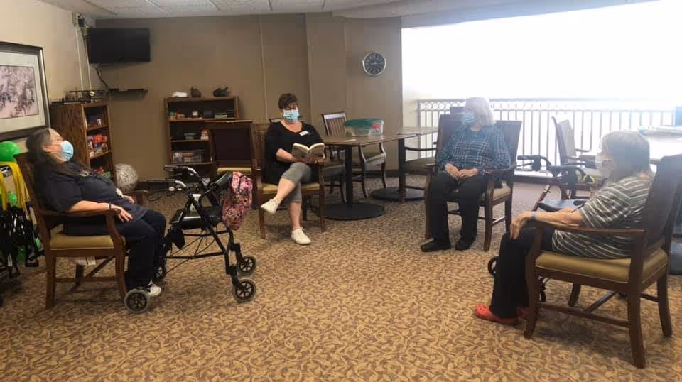 Four elderly women wearing face masks sitting in a circle on chairs in a carpeted room. One woman is reading a book while the others listen. There is a walker next to one of the women, a bookshelf with various items, a wall clock, and a large window with bright light coming through.