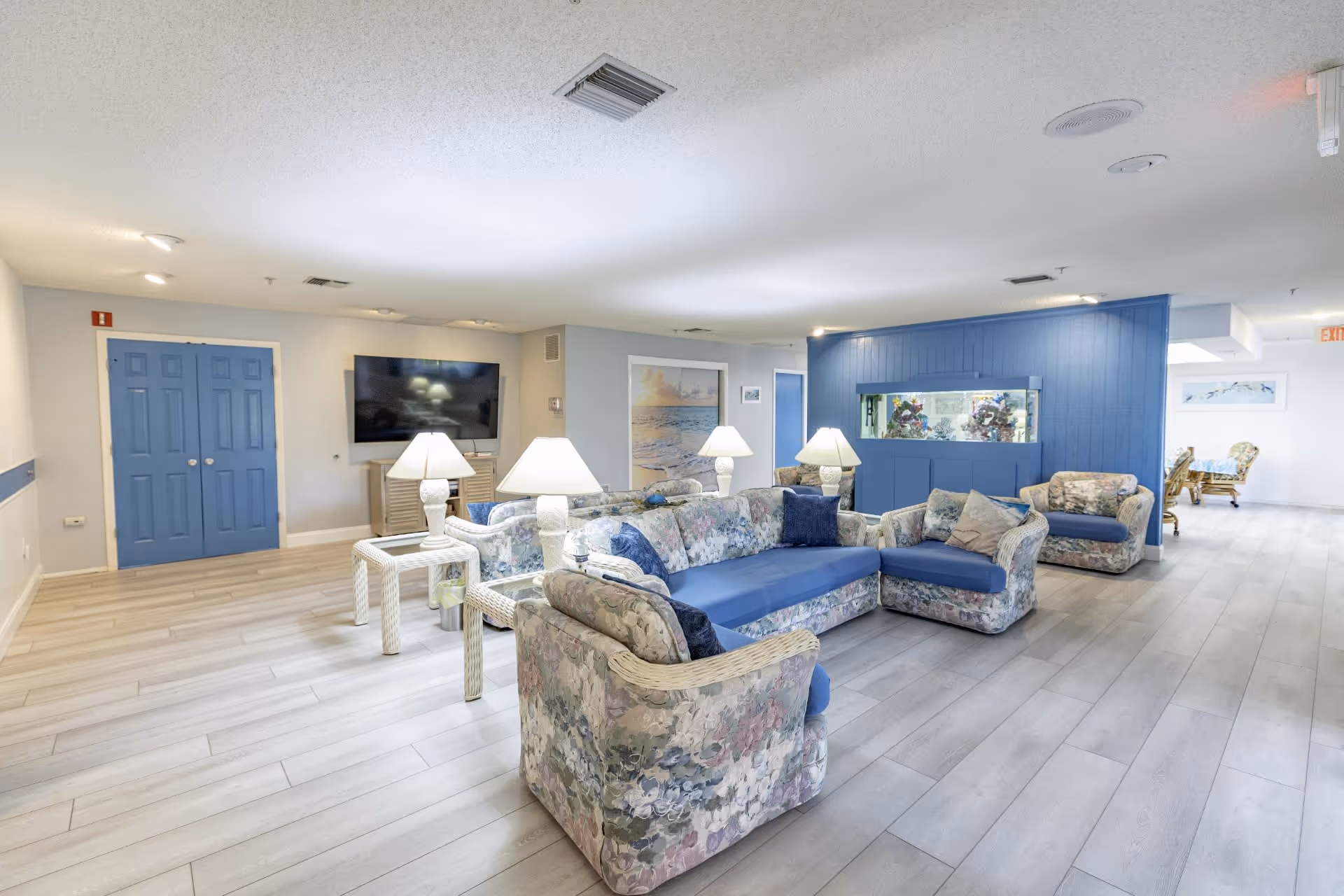 A spacious living room area in an assisted living facility featuring floral-patterned sofas with blue cushions, white side tables with lamps, a large flat-screen TV mounted on the wall, and a blue accent wall with a built-in aquarium. The room has light wood flooring and soft lighting, with a dining area visible in the background.