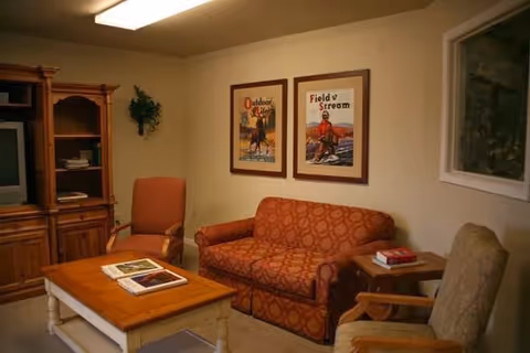 A cozy living room with a patterned orange sofa, two armchairs, a wooden coffee table with books on it, a wooden cabinet with shelves, and two framed vintage-style posters on the wall.