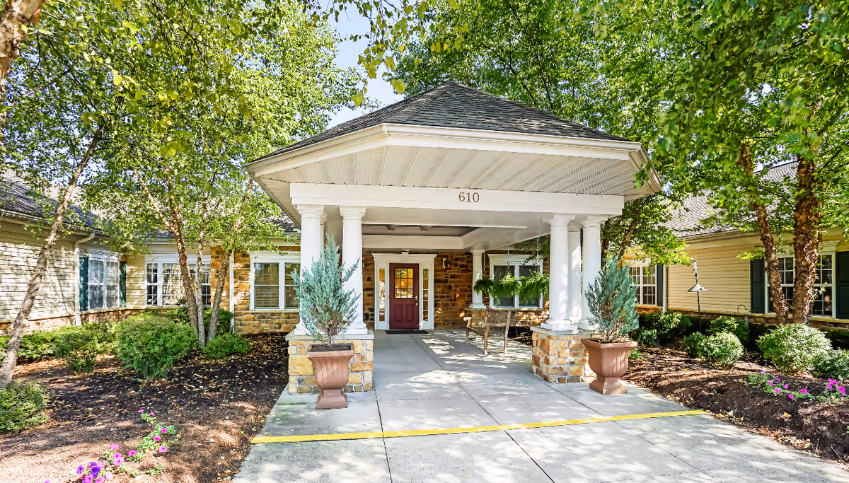 Entrance of a senior living facility with a covered driveway supported by white columns. The building has stone and beige siding with multiple windows. There are potted plants and trees around the entrance, and a bench is visible under the covered area. The address number 610 is displayed above the entrance.
