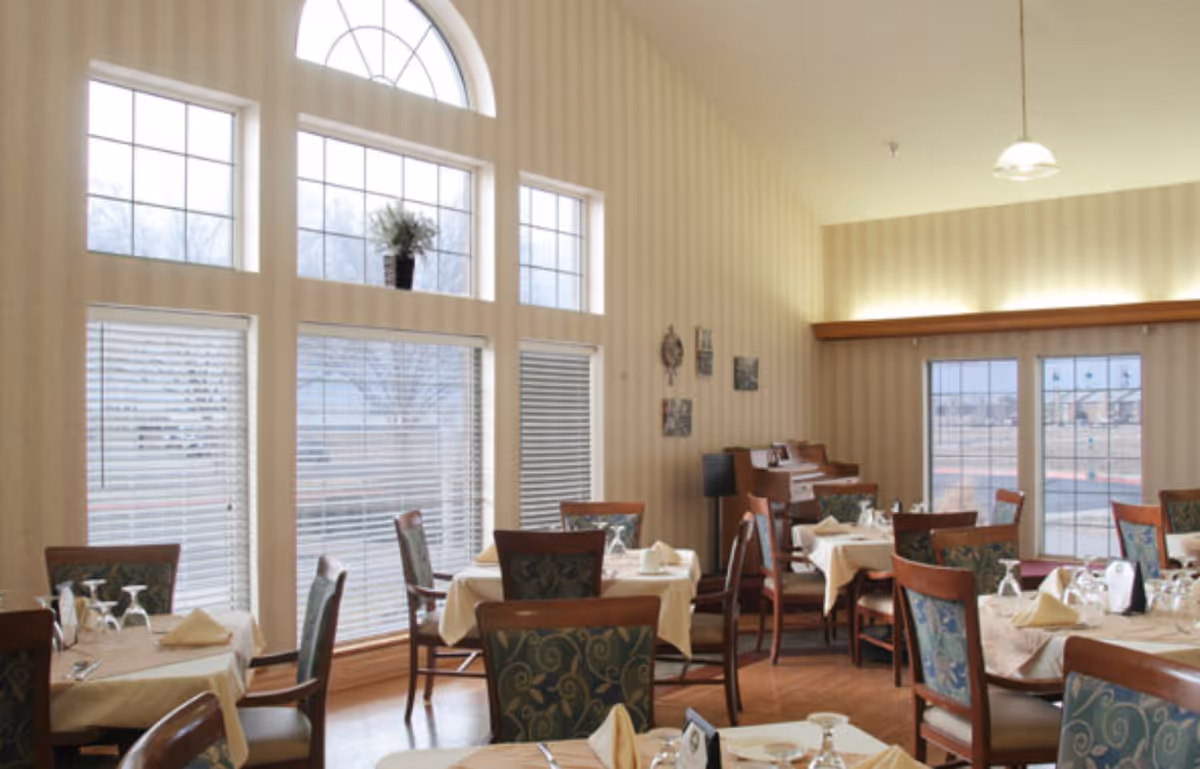 Dining room with several tables covered with beige tablecloths, set with napkins and glassware. The room has large windows with blinds and an arched window above, letting in natural light. There is a piano against the far wall and framed pictures hanging on the striped wallpaper. Wooden chairs with patterned upholstery surround the tables.