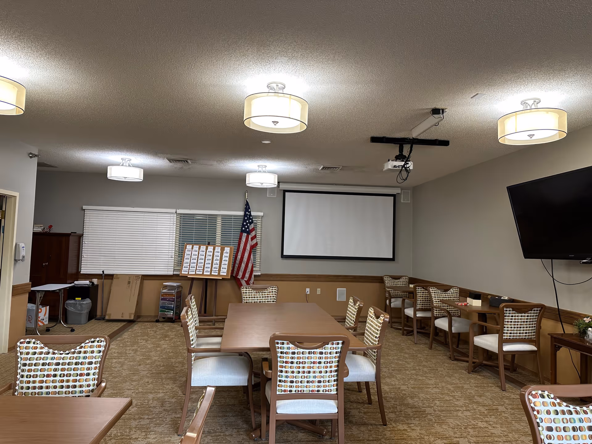 A meeting or activity room with several wooden tables and chairs featuring patterned upholstery. The room has beige walls and carpet, ceiling lights, a large flat-screen TV mounted on the right wall, and a projector with a screen at the front. An American flag stands near the window, and a board with numbers is displayed on an easel.