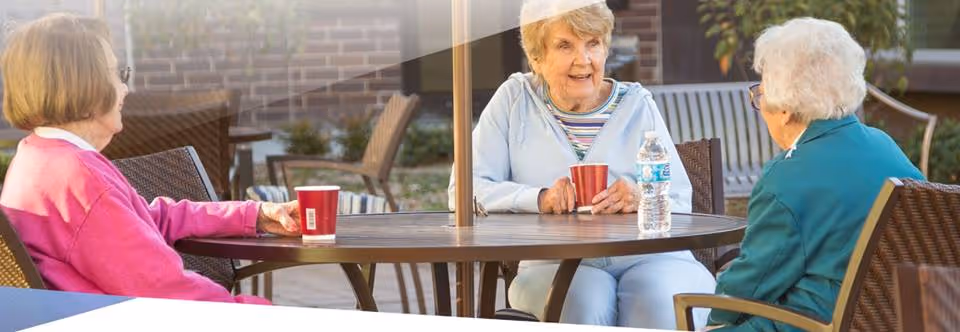 Three elderly women sitting around a round outdoor table having a conversation. Two of them have red cups in front of them, and one has a water bottle. They are seated on patio chairs with a brick wall and greenery in the background.