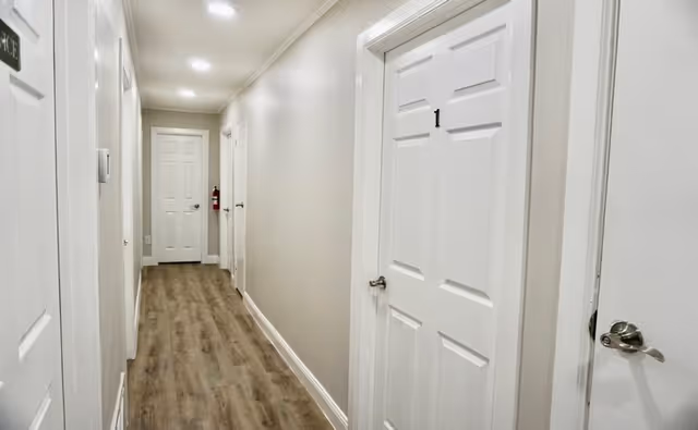 Well-lit interior hallway with white numbered doors and wood-look flooring.