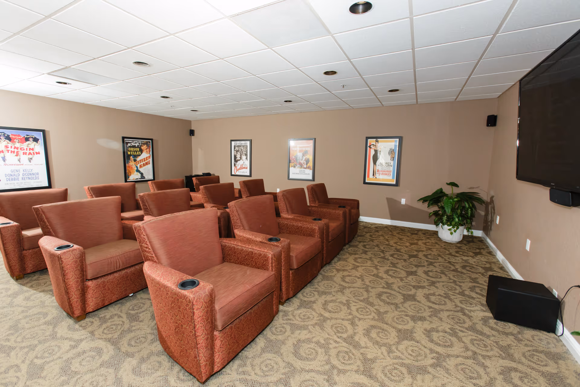 A small movie theater room with two rows of red cushioned armchairs, each with a cup holder. The walls are painted beige and decorated with framed classic movie posters. A large flat-screen TV is mounted on the right wall above a black speaker. There is a potted plant in the corner of the room and patterned carpet on the floor.