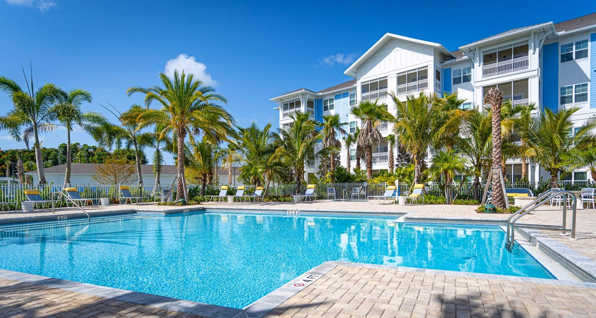 Outdoor swimming pool with clear blue water surrounded by palm trees and lounge chairs, with a multi-story residential building in the background under a bright blue sky.