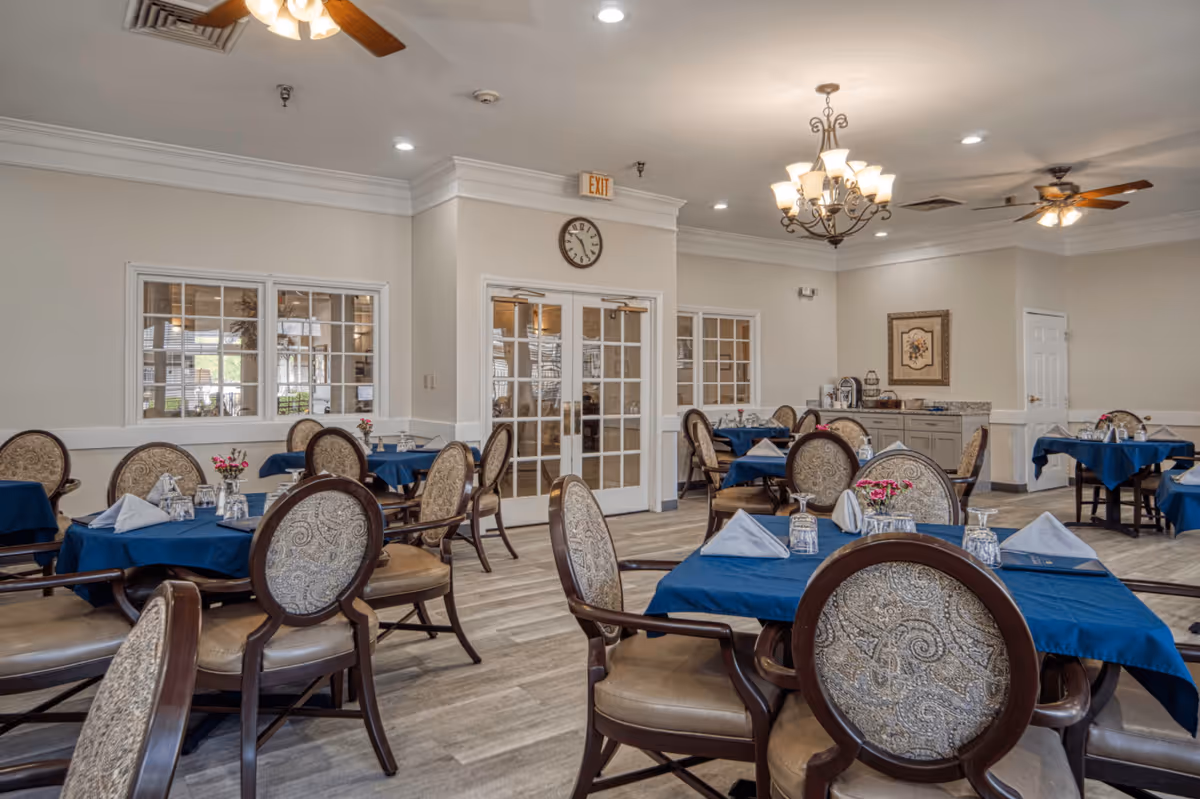 A dining room with multiple tables covered in blue tablecloths, each set with napkins, glasses, and small flower arrangements. The room features upholstered chairs with patterned backs, ceiling fans, a chandelier, and a clock above double glass doors. The walls are light-colored with white trim, and there is a sideboard with a coffee station and framed artwork.