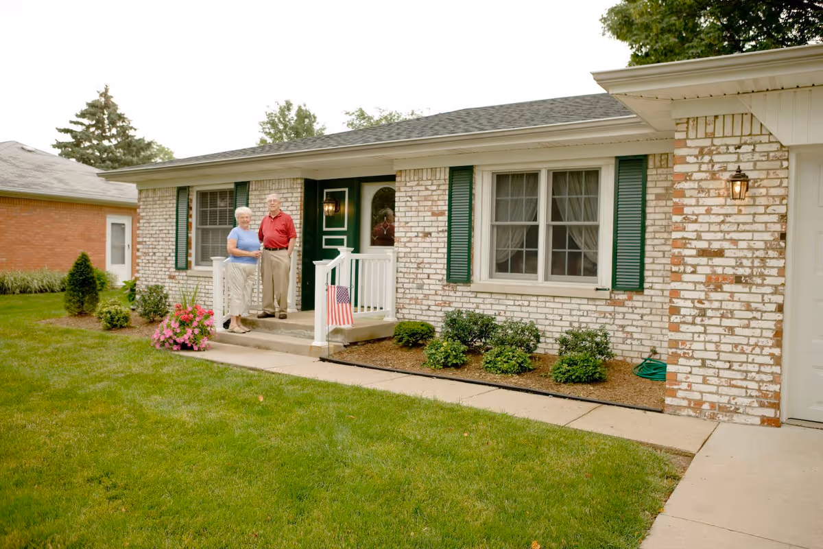 An elderly couple standing on the front porch of a single-story brick house with green shutters and a green door. The porch has white railings and an American flag displayed. There are small bushes and flowers planted along the front of the house, and a well-maintained lawn in the foreground.