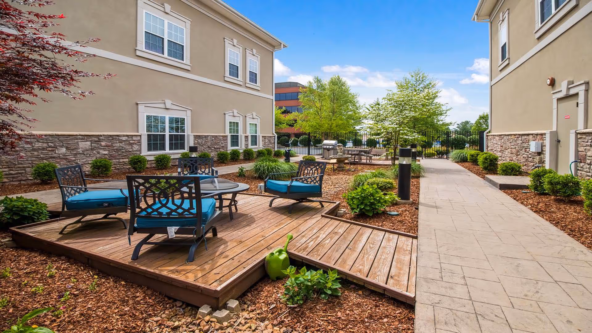 Outdoor patio area between two beige buildings with stone accents, featuring a wooden deck with a glass-top table and four chairs with blue cushions. The area is landscaped with small bushes, trees, and mulch, and includes a green watering can and a barbecue grill in the background under a partly cloudy sky.