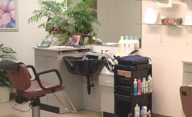Interior view of a hair salon area within a facility, featuring a brown salon chair, a black hair washing sink, a rolling cart with various hair care products, and shelves with additional bottles and items. There are plants and framed pictures on the walls, creating a welcoming atmosphere.