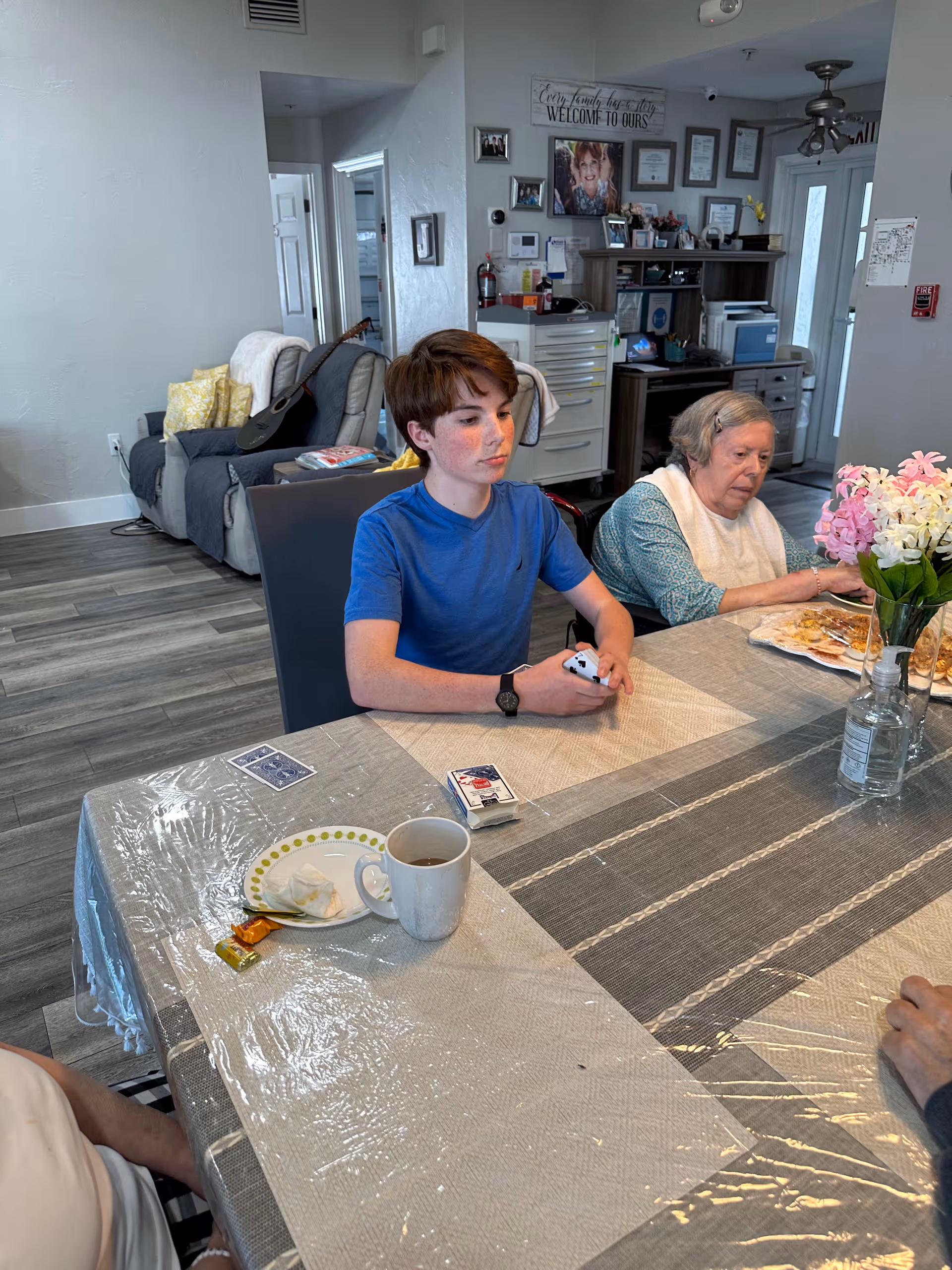 Two people sit at a dining table in a senior living facility's communal room with cups, plates, playing cards, and a vase of flowers on the table.
