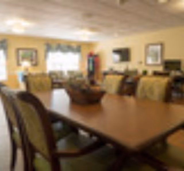 A dining room with a large wooden table surrounded by cushioned chairs. The room has warm lighting, windows with curtains, framed pictures on the walls, and a cozy atmosphere.