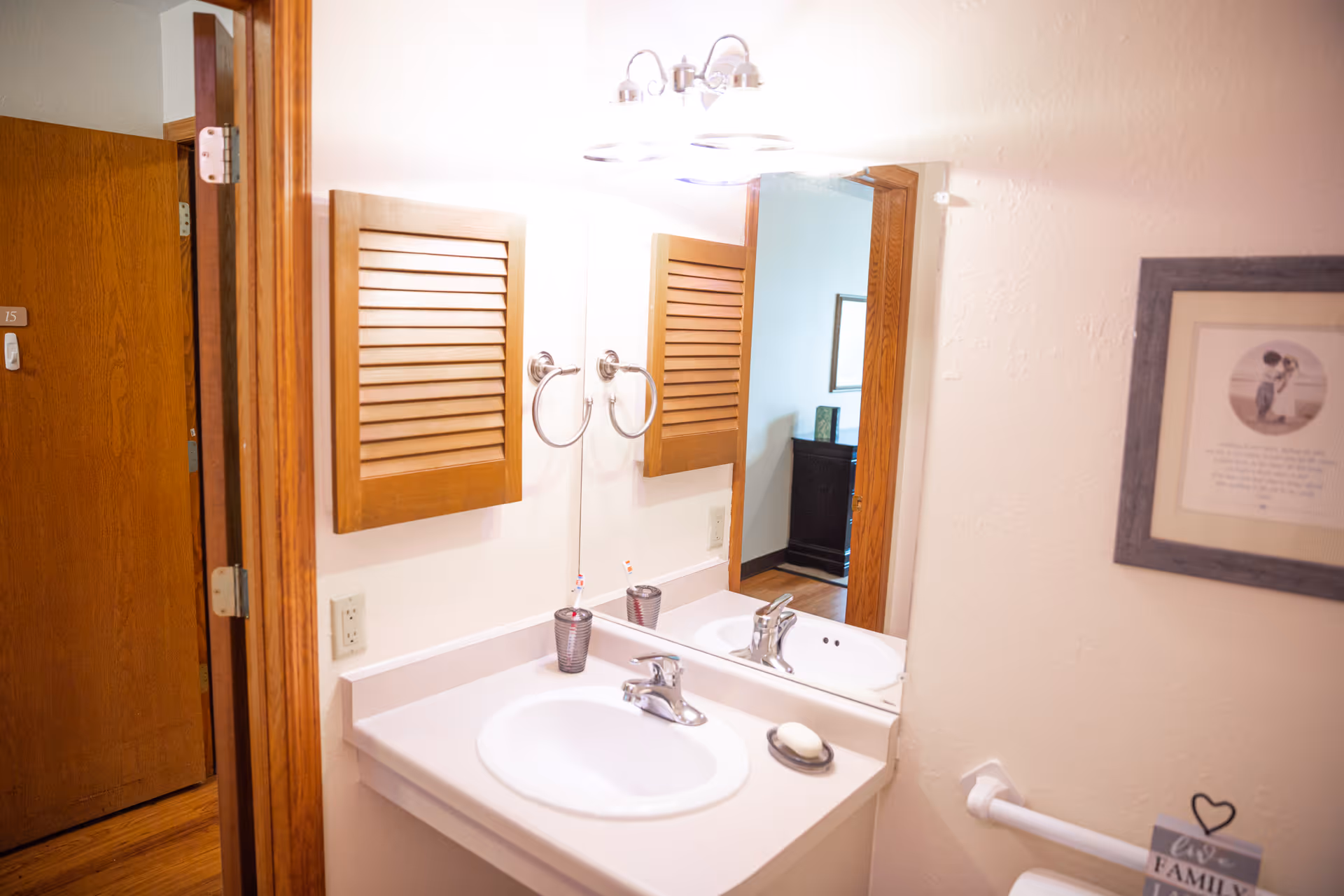 Bathroom vanity with a sink, mirror, wooden louvered medicine cabinet, towel ring and a doorway to an adjacent room.