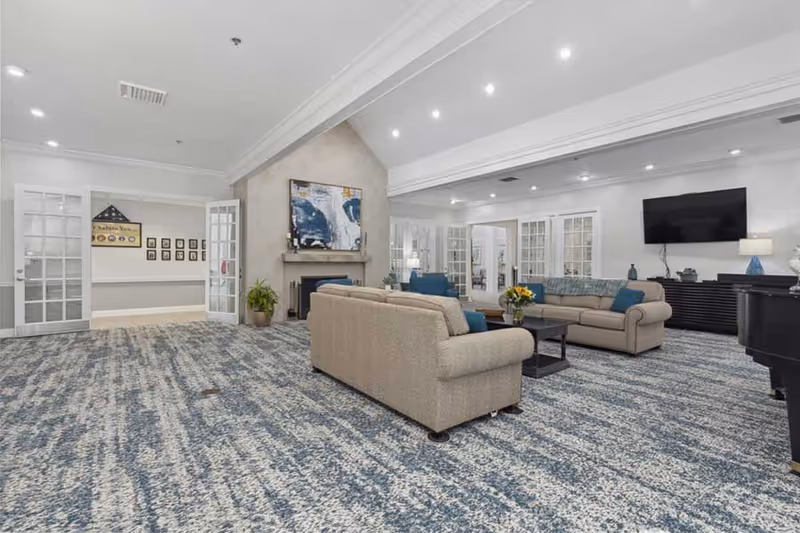 A spacious living room area with two beige sofas and a coffee table in the center. The room features a patterned blue and gray carpet, a fireplace with artwork above it, a large flat-screen TV mounted on the wall, and multiple glass-paneled doors leading to other rooms. The ceiling has recessed lighting and the walls are painted white.