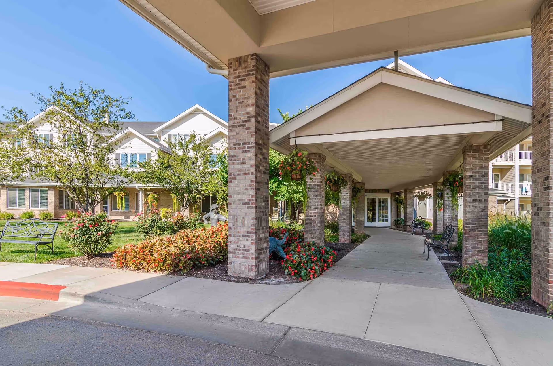 Covered entrance walkway to a senior living facility with brick pillars, hanging flower baskets, benches along the path, and landscaped garden beds with flowers and shrubs. The building has multiple windows and a light-colored exterior under a clear blue sky.