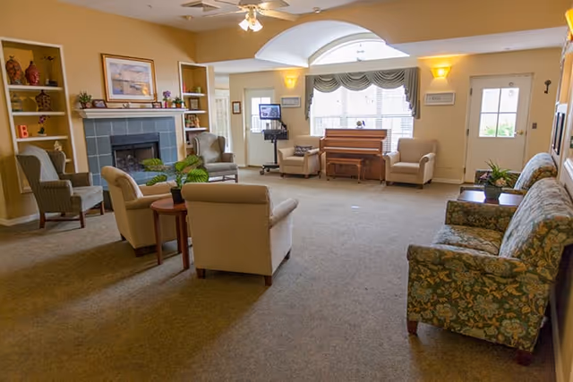 A spacious living room area with multiple upholstered chairs and sofas arranged around a central space. There is a fireplace with decorative items on the mantel, a piano near a large window with drapes, and a door leading outside. The room is warmly lit with wall sconces and a ceiling fan with lights.