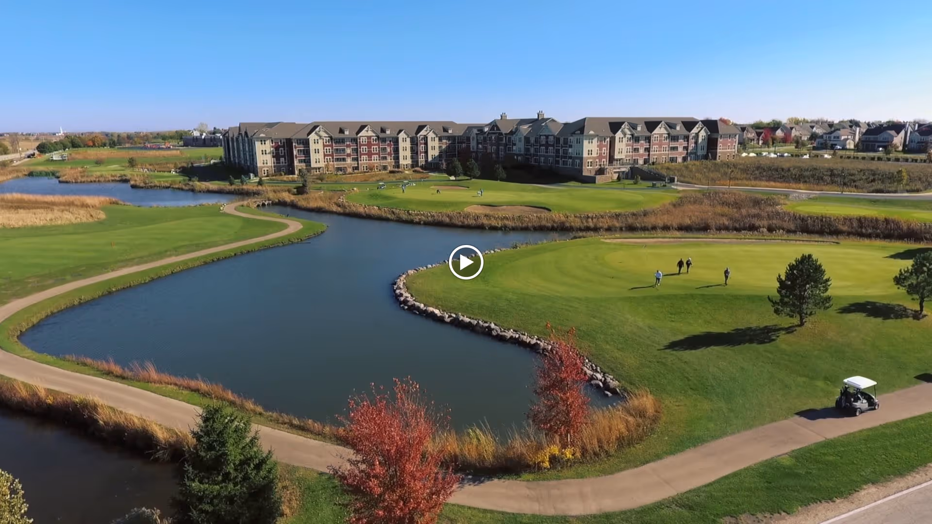 A large senior living facility building situated behind a golf course with green fairways, a water hazard, and a paved golf cart path. Several people are seen walking on the golf course, and a golf cart is visible on the path. The sky is clear and blue.