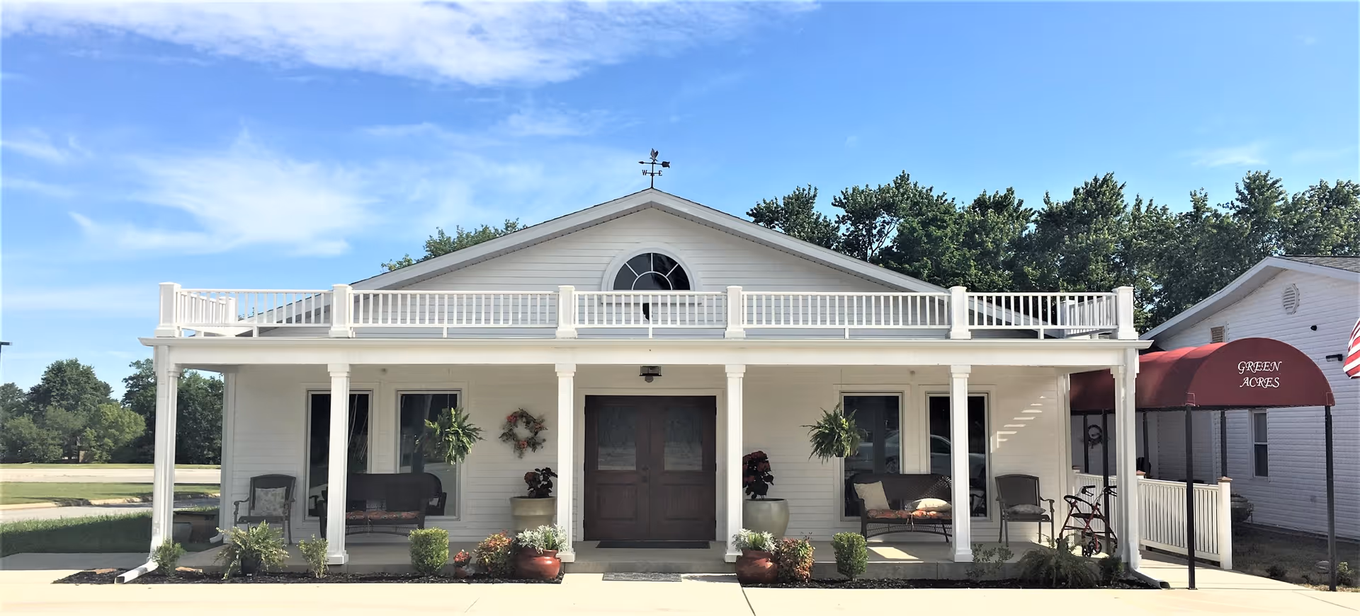 Front exterior of a white single-story building with a covered porch supported by columns, potted plants and seating, and a red awning reading 'GREEN ACRES'.