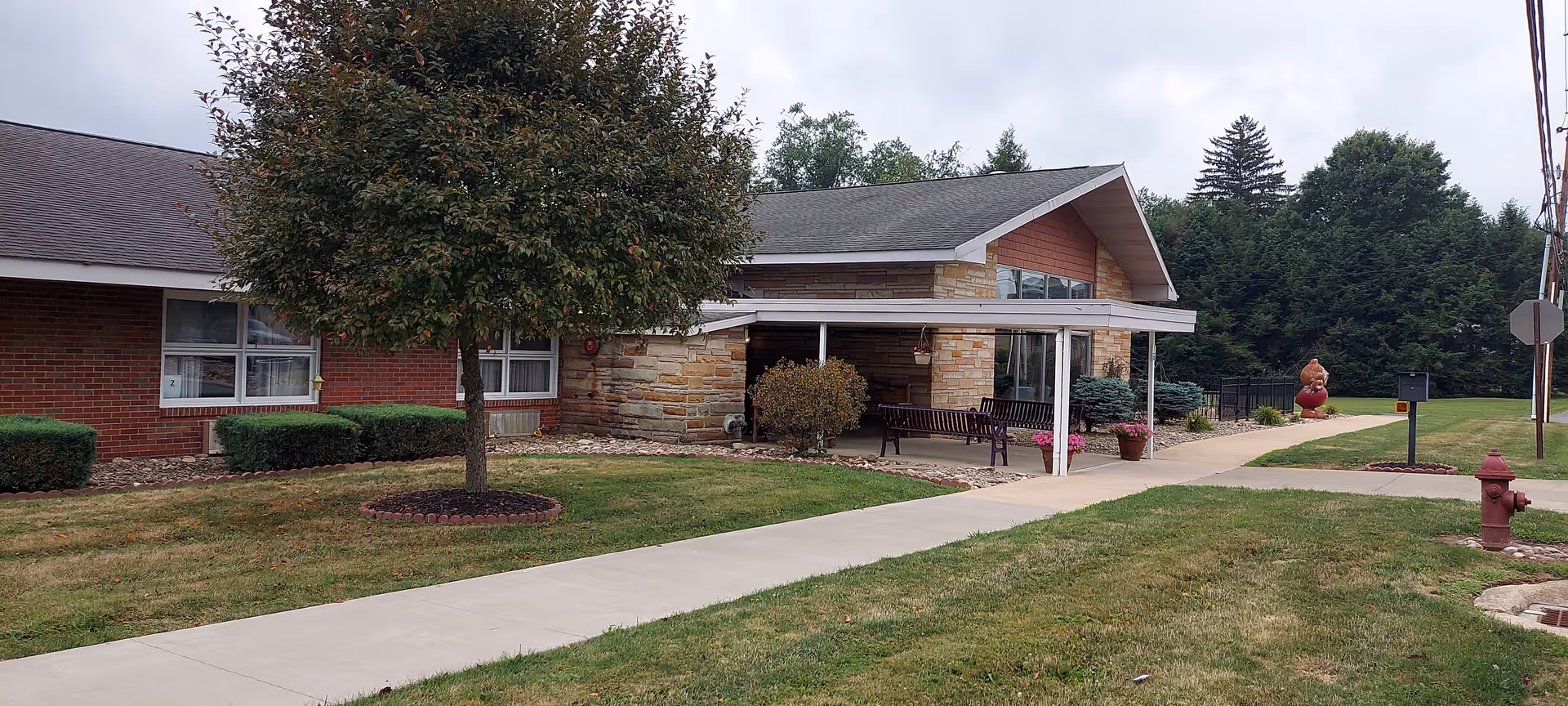 Exterior view of Mulberry Healthcare and Rehabilitation Center showing a single-story building with a brick and stone facade, a covered entrance with benches, a tree, and a sidewalk leading to the entrance. There is a fire hydrant and some landscaping with grass and bushes.