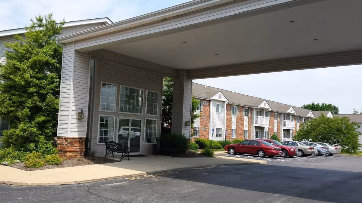 Exterior view of Heritage Place senior living facility showing the main entrance with a covered drop-off area, a bench near the entrance, and a parking lot with several parked cars. The building has a combination of brick and siding with multiple windows and small balconies.