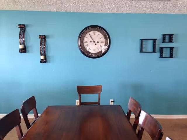 A dining room with a wooden table surrounded by six wooden chairs. The wall behind the table is painted teal and features a large round clock in the center, two decorative candle holders on the left, and three small black wall shelves on the right.