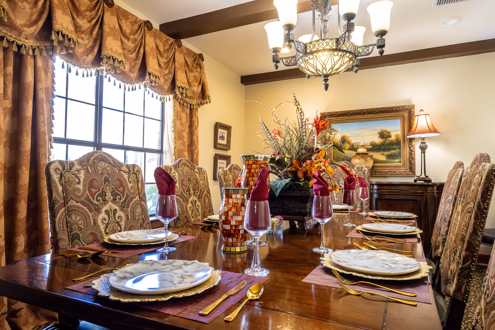 Elegant dining room with a polished wooden table set for six, featuring patterned upholstered chairs, gold-rimmed plates, gold cutlery, wine glasses with folded burgundy napkins, a decorative centerpiece with flowers, and warm lighting from a chandelier and table lamp. Large windows with ornate curtains allow natural light into the room.