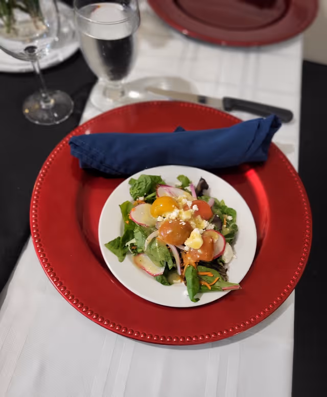 A small mixed salad on a white plate set on a red charger with a rolled navy napkin and glassware on a white tablecloth.
