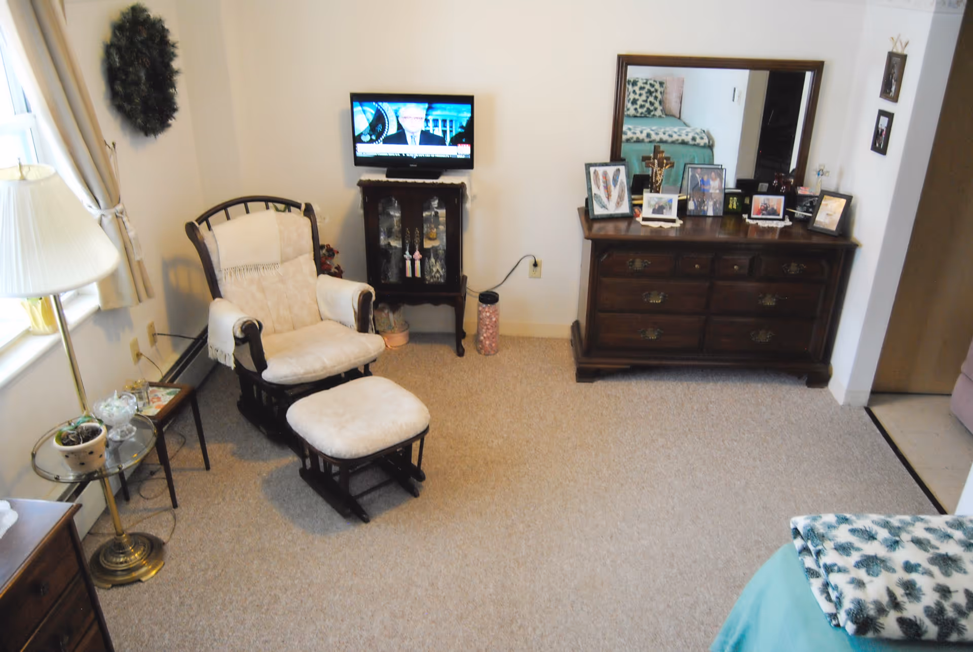 Cozy bedroom sitting area with an upholstered rocking chair and ottoman, a dresser with mirror, small TV, and a bed partially visible.