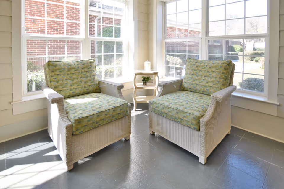 Two cushioned wicker armchairs with green patterned cushions are placed in a sunlit corner with large windows showing an outdoor view of bushes and a brick building. A small wooden side table with a plant and a cup is positioned between the chairs.