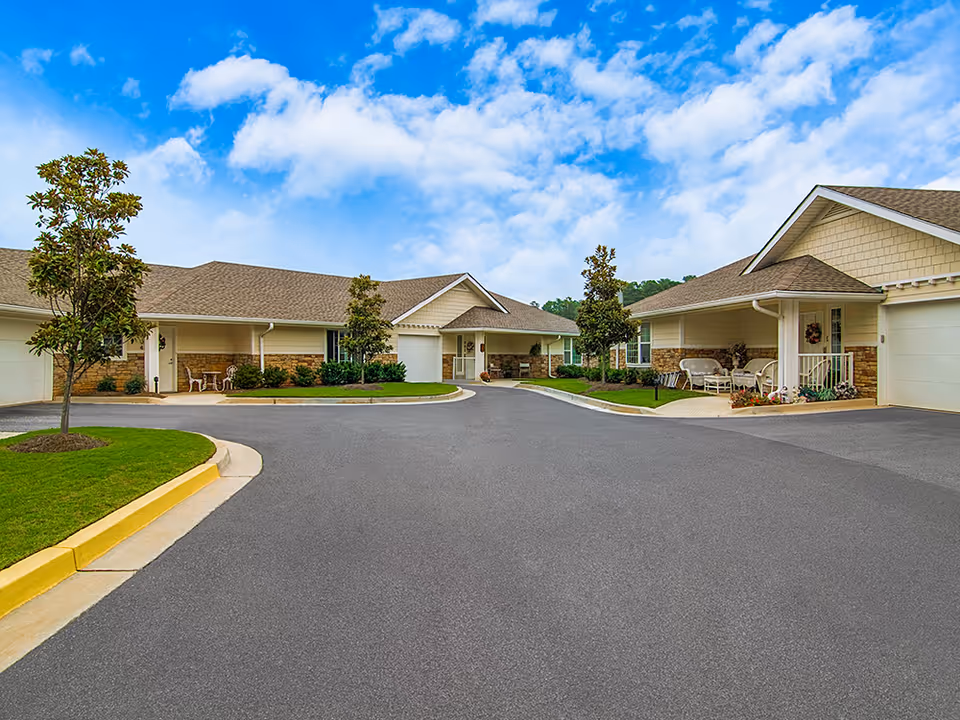 Exterior view of Laurel Grove by Barclay House showing a paved driveway surrounded by single-story buildings with beige siding and stone accents, small trees, and a bright blue sky with scattered clouds.