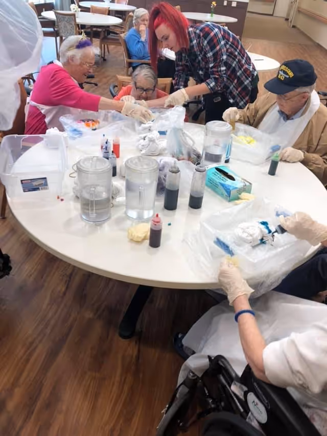 A group of elderly people and a caregiver with red hair gathered around a round table engaging in a craft activity involving tie-dye materials. The table has various bottles of dye, water pitchers, gloves, and plastic coverings. The setting appears to be a communal area with wooden flooring and other tables and chairs in the background.