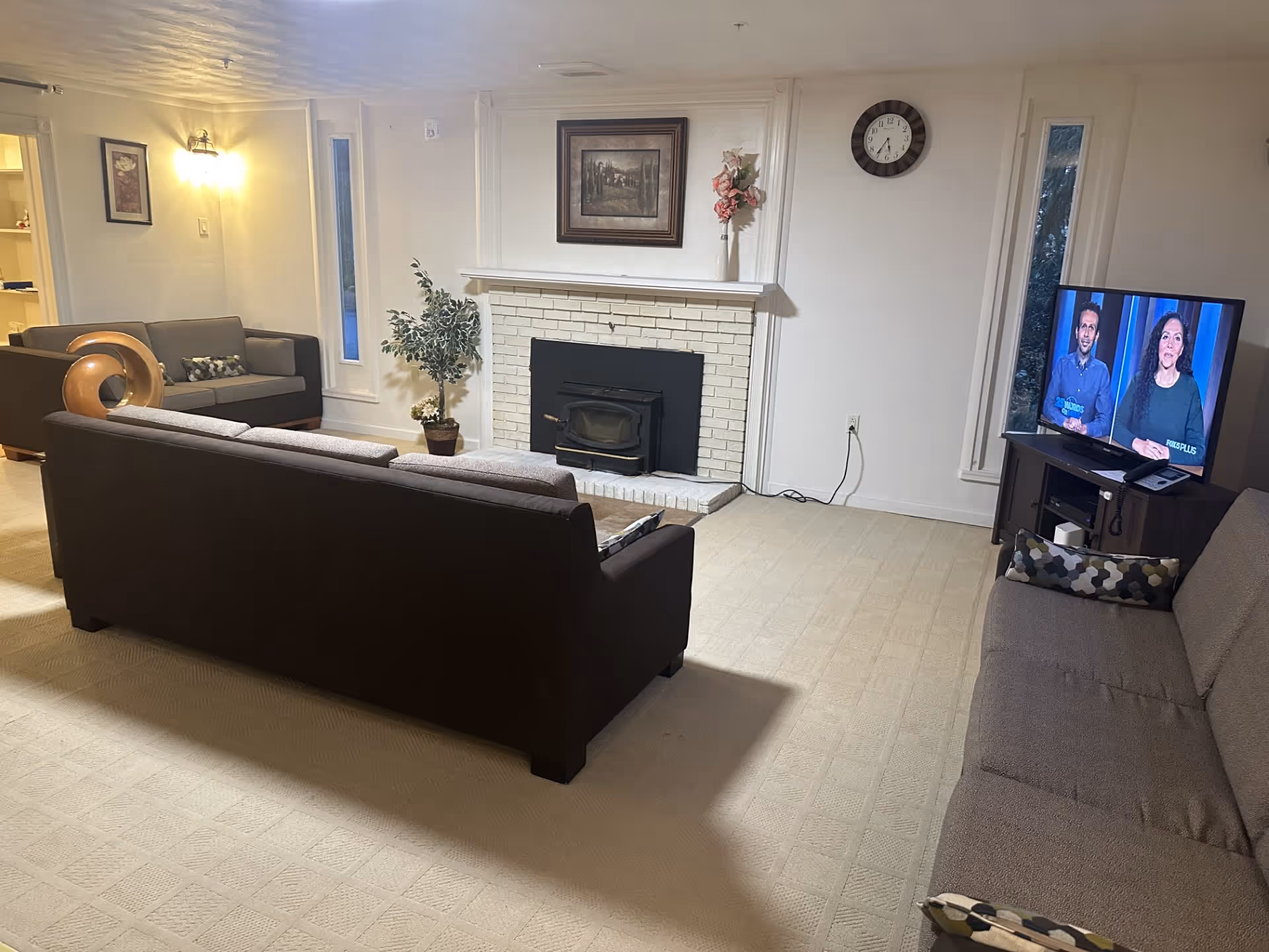 A cozy living room with three gray sofas arranged around a white brick fireplace. There is a potted plant and a vase with flowers on the mantel. A wall clock is mounted above the fireplace. A television on a wooden stand is showing two people speaking. The room has beige carpet and soft lighting.