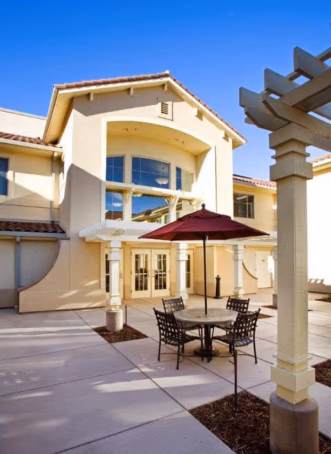 Outdoor patio area at Westmont of Brentwood with a round table, four metal chairs, and a red umbrella. The patio is paved and adjacent to a beige building with large windows and a covered entrance. A pergola structure is visible on the right side of the image under a clear blue sky.