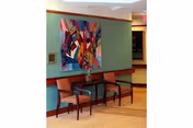 A seating area with two wooden chairs with orange cushions and a small dark wooden table between them, placed against a green wall. Above the chairs, there is a colorful abstract painting. The floor has a light brown carpet, and there is a handrail along the wall.