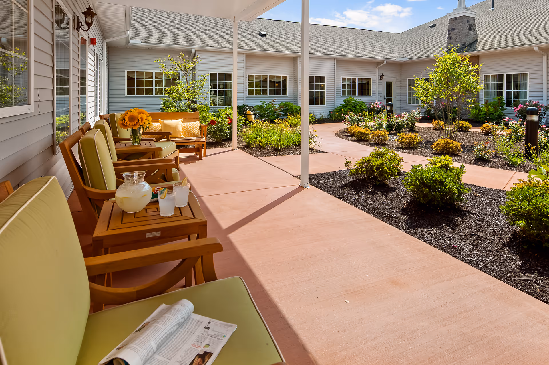 Covered courtyard patio at a senior living facility with cushioned chairs, a table with a pitcher of lemonade and sunflowers, and landscaped garden beds.